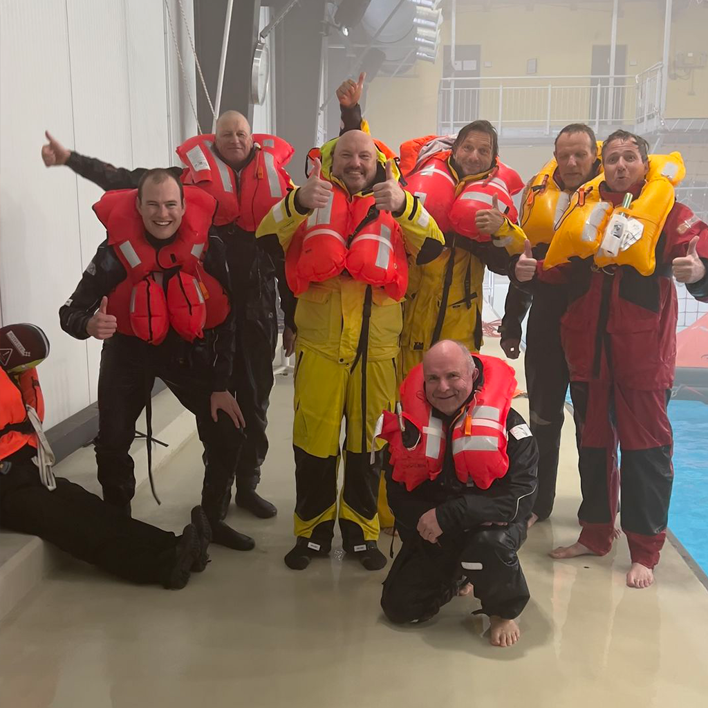Teilnehmergruppe am Beckenrand mit aufgeblasenen Rettungswesten beim Offshore-Sicherheitstraining, lachend und motiviert