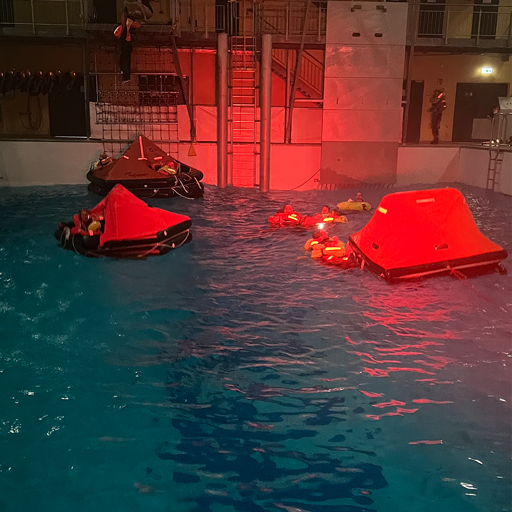 Großaufnahme des Offshore-Trainingsbeckens mit parallelen Übungen: Helikopterbergung, Rettungsinsel und Schwimmen mit Rettungswesten im Wasser