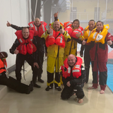 Teilnehmergruppe am Beckenrand mit aufgeblasenen Rettungswesten beim Offshore-Sicherheitstraining, lachend und motiviert